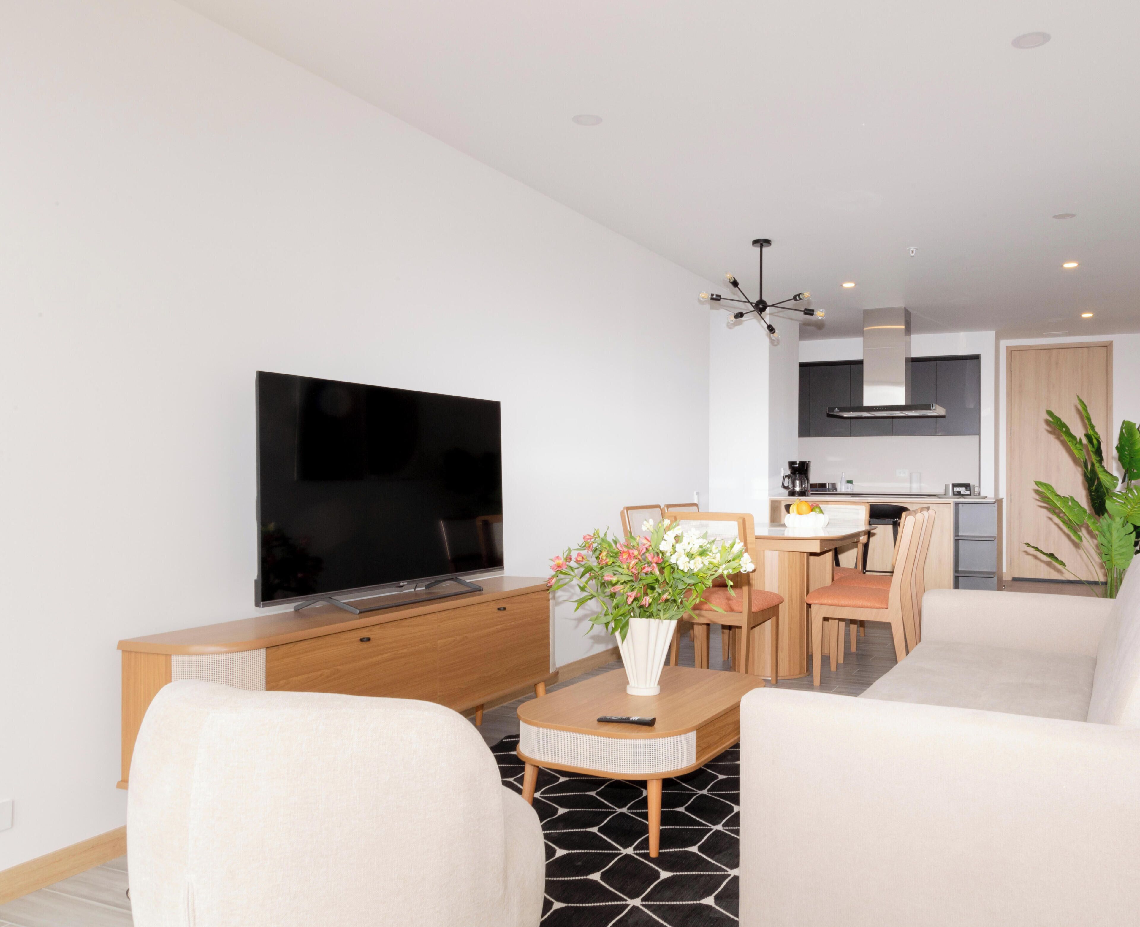 Een moderne woonkamer met een flatscreen televisie, een bank, een eettafel met stoelen en een keuken in de achtergrond.