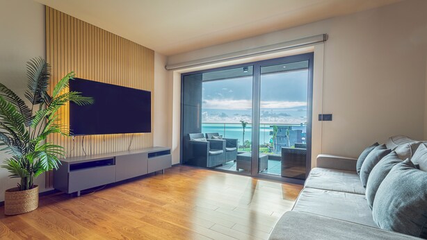 Un salon moderne doté d’un téléviseur à écran plat, de parquet, d’un canapé gris et d’une grande porte-fenêtre coulissante donnant sur un balcon avec vue sur la mer.