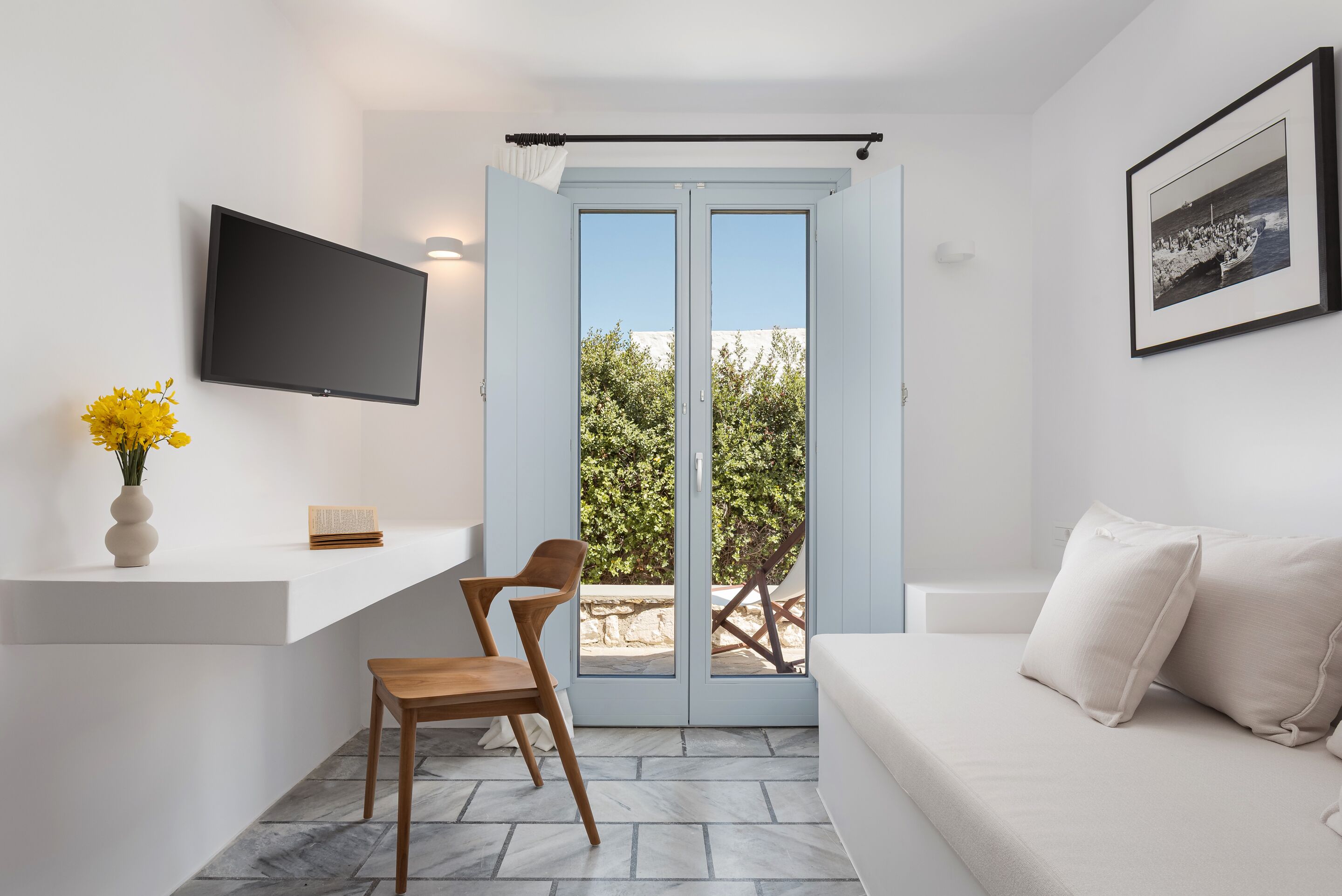 Une chambre moderne équipée d’un téléviseur à écran plat, d’une chaise en bois, d’un canapé, d’une petite table avec un vase, et d’une porte donnant sur un balcon avec vue sur des arbres et un immeuble.