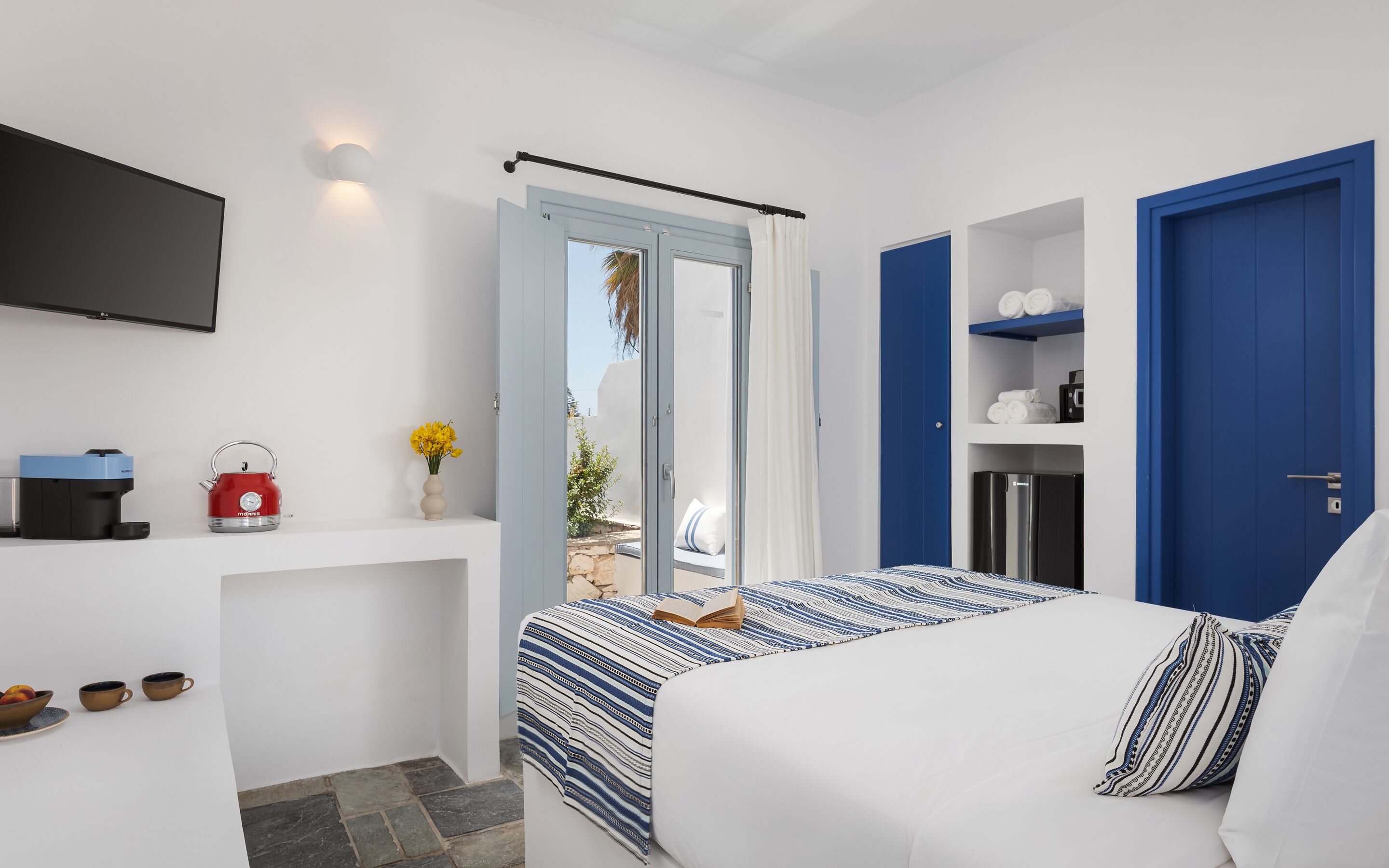 Una habitación de hotel moderna con una cama, un televisor de pantalla plana, una cafetera y una mesita con tazas y fruta.