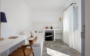 A modern, minimalist room with a stone floor, a wooden chair, a white desk, a small kitchenette, and a bathroom with a shower curtain. - Soleil Hotel Paros (Drios)