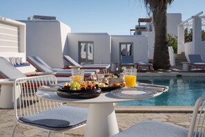 Un área de comedor junto a la piscina con una mesa dispuesta para el desayuno, incluyendo frutas, huevos y jugo, con vistas a una piscina con sillas de descanso y un edificio blanco al fondo.