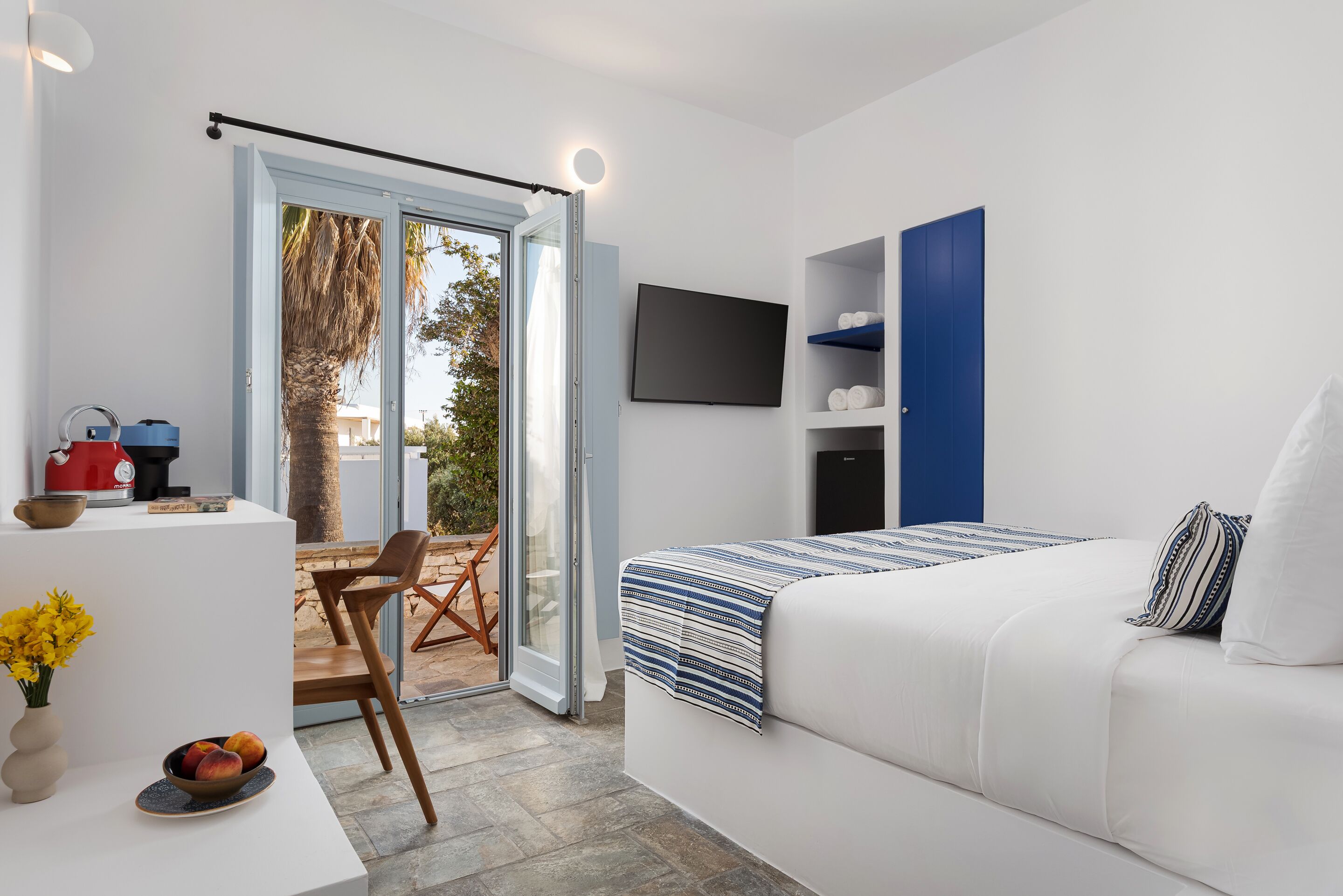 A modern bedroom with a bed, a TV, a desk with a coffee machine, and a sliding glass door to an outdoor area.
