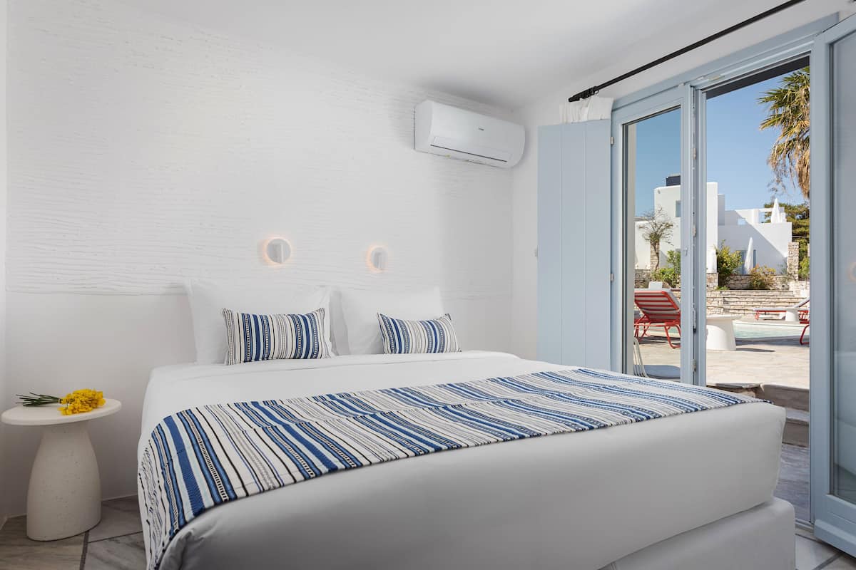 Una habitación de hotel moderna con una cama grande, una mesita de noche pequeña con un florero y un equipo de aire acondicionado integrado, además de un ventanal corredero que da a una zona de piscina.