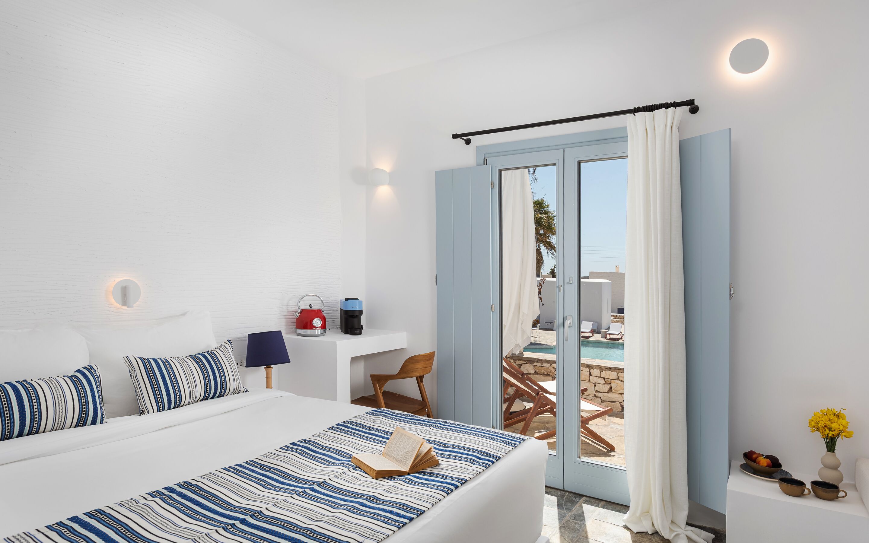 A hotel room with a bed, a desk, a chair, a coffee machine, a kettle, a table with chairs outside, and a view of the pool.