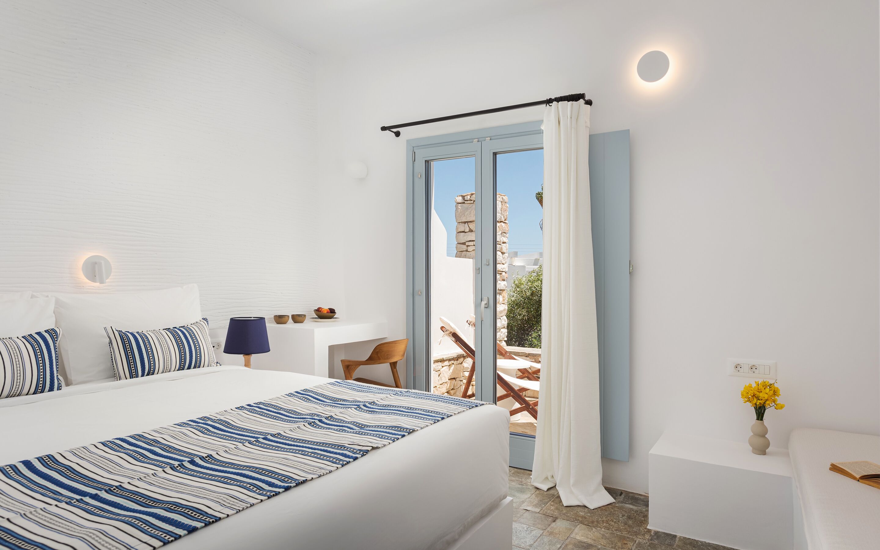 A bedroom with a bed, a nightstand, a lamp, a window with curtains, and a view of the outside.