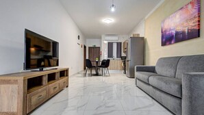 Un salon moderne avec un téléviseur à écran plat, un canapé gris, un meuble de rangement en bois, un coin repas avec des chaises et une cuisine en arrière-plan.