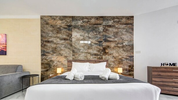 Une chambre d’hôtel moderne avec un grand lit, un canapé gris, une commode en bois et un mur en pierre.