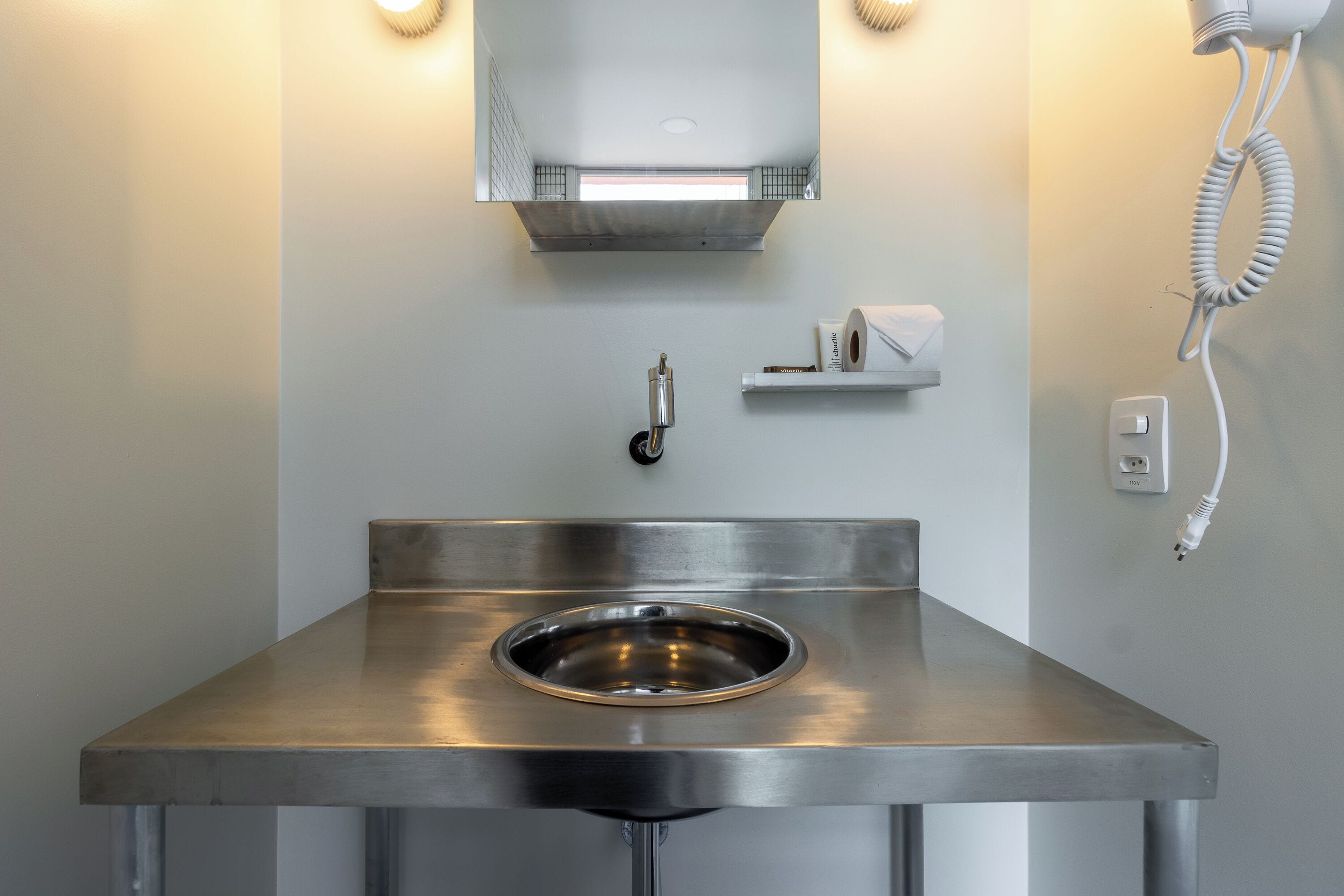 Un baño moderno con lavamanos de acero inoxidable, un espejo, un secador de pelo y un dispensador de toallas.