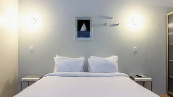 Une chambre d’hôtel comprenant un lit, deux tables de chevet, une petite table, un luminaire fixé au mur et un tableau encadré.