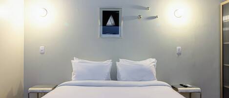 A hotel room with a bed, two bedside tables, a nightstand, a wall-mounted light fixture, and a framed picture.