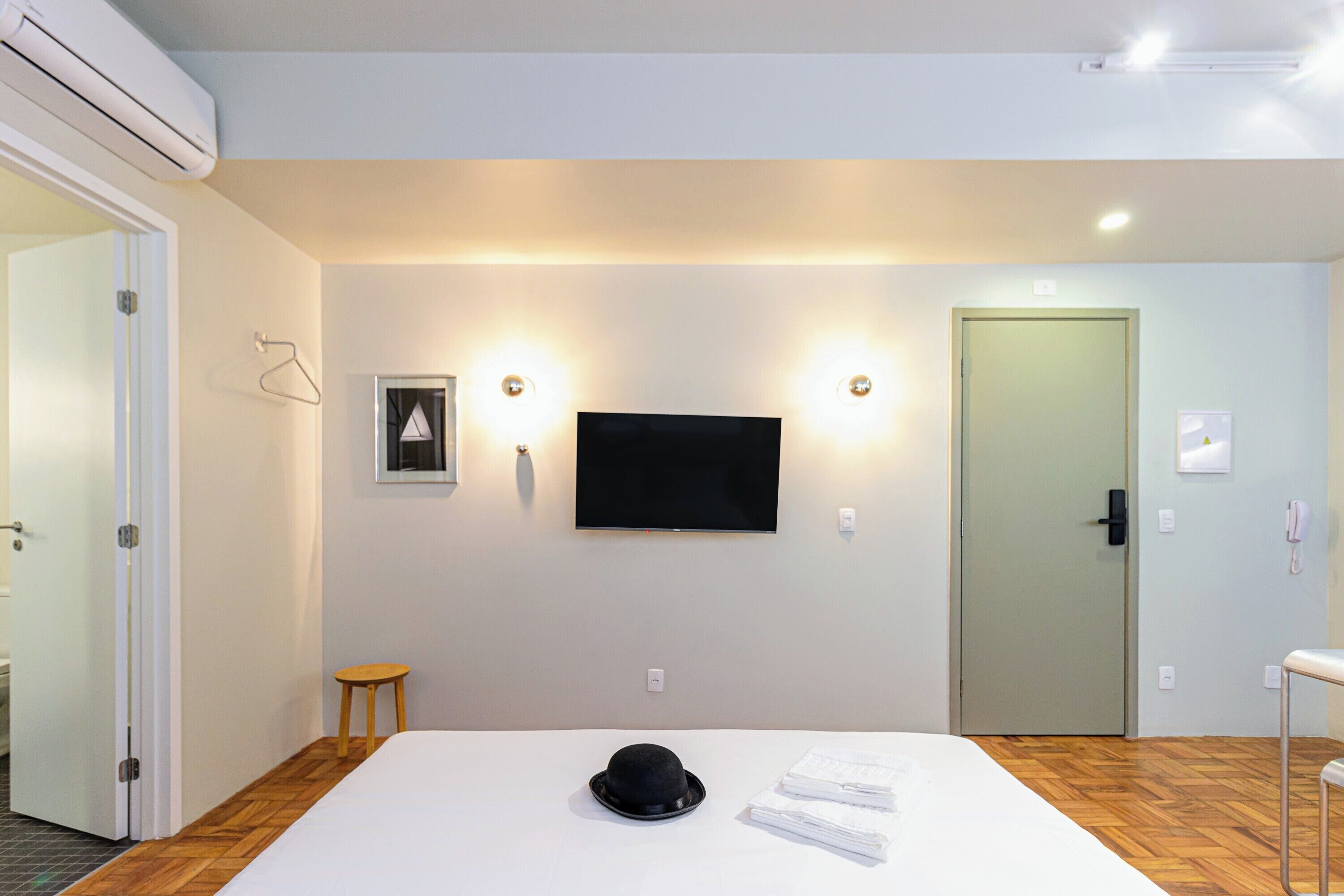 Hotellihuone, jossa on sänky, seinälle asennettu televisio, ovi ja pieni jakkara.