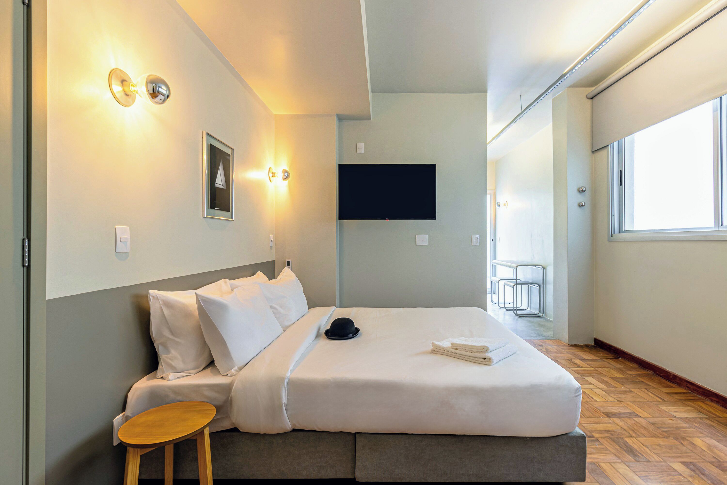 A modern hotel room with a bed, bedside table, wall-mounted TV, and a window.