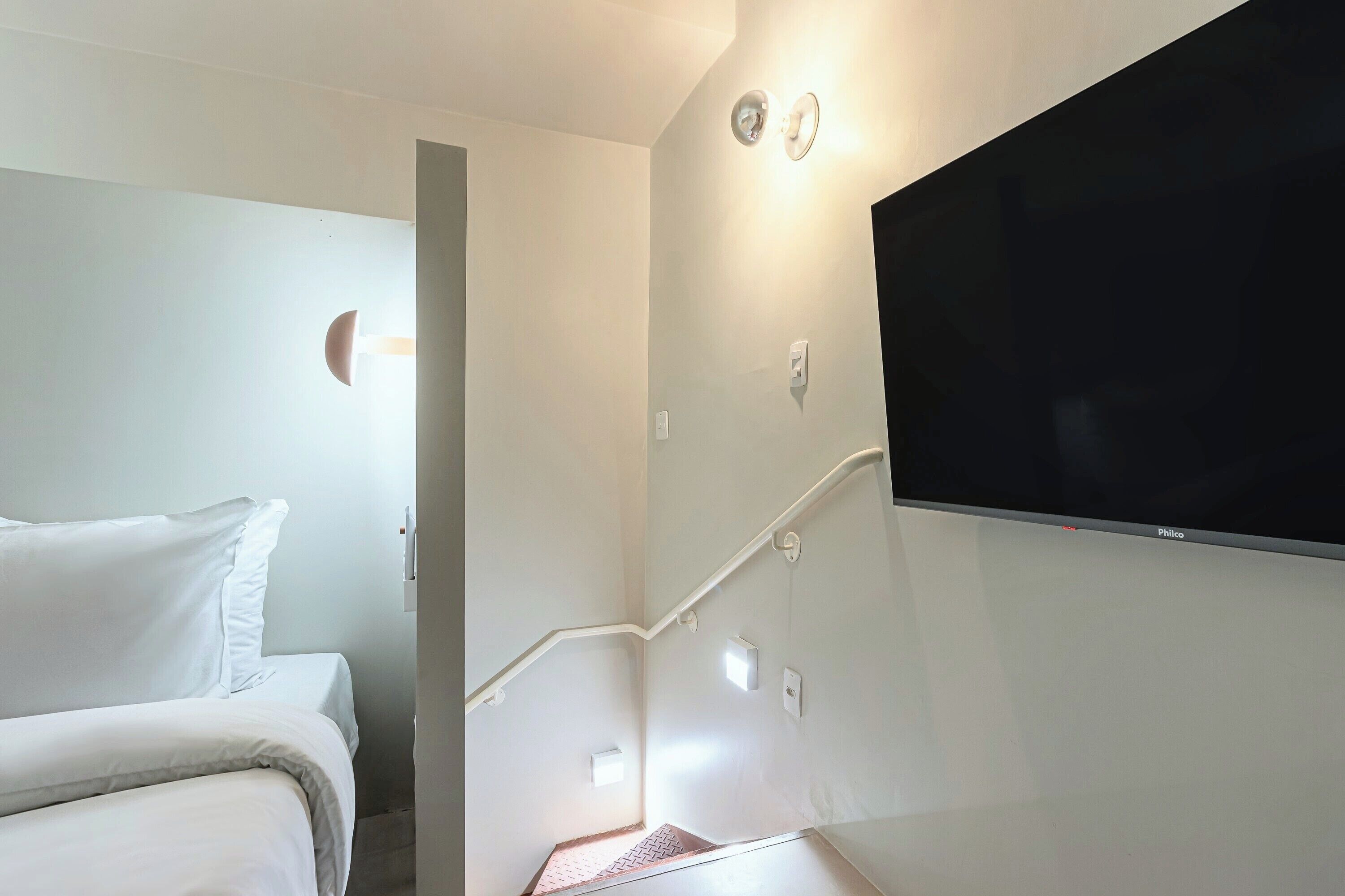 A hotel room with a bed, a flat-screen TV mounted on the wall, a staircase, and a small window.