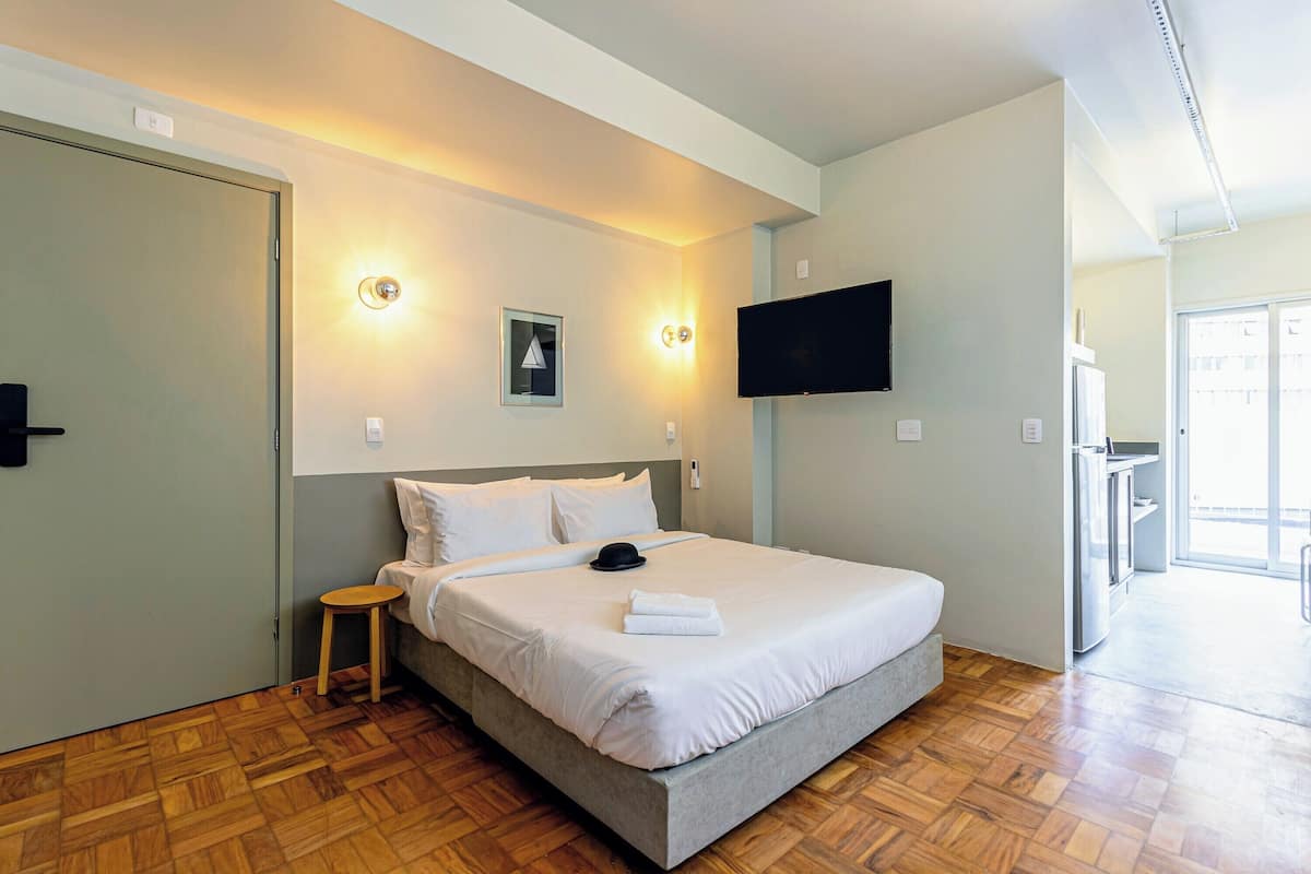 Een hotelkamer met een bed, een aan de muur bevestigde televisie, een klein krukje en een deur.
