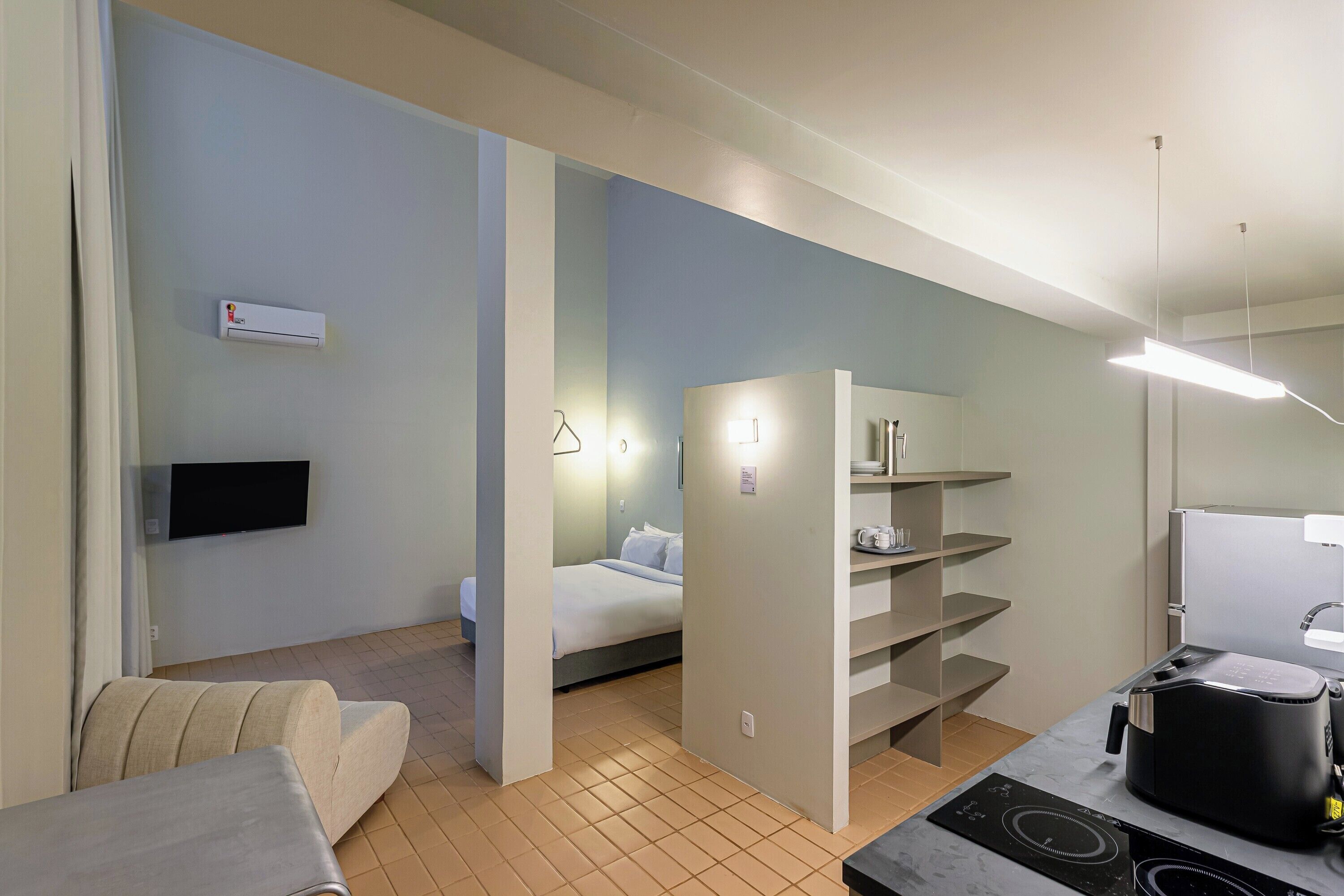 Quarto de hotel moderno com cama, sofá, TV de tela plana, cozinha compacta com fogão e estante embutida.