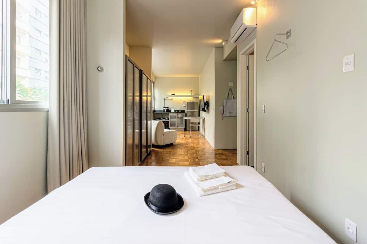 Hotelkamer met een bed, een zwarte hoed op het bed, een keuken in de achtergrond en een raam met gordijnen.