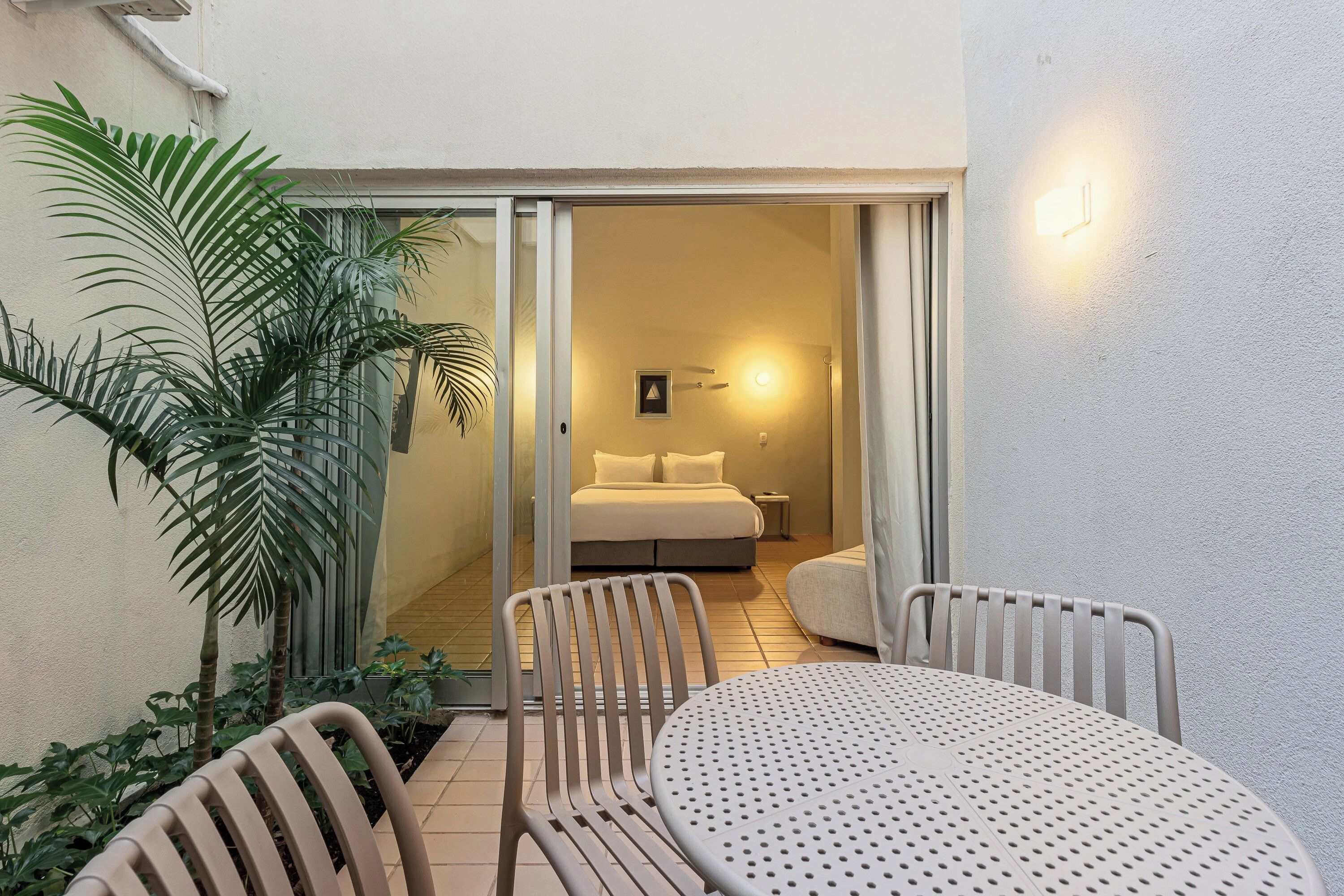 A room with a bed, a small table, and chairs, a sliding glass door leading to another room, and a potted plant.