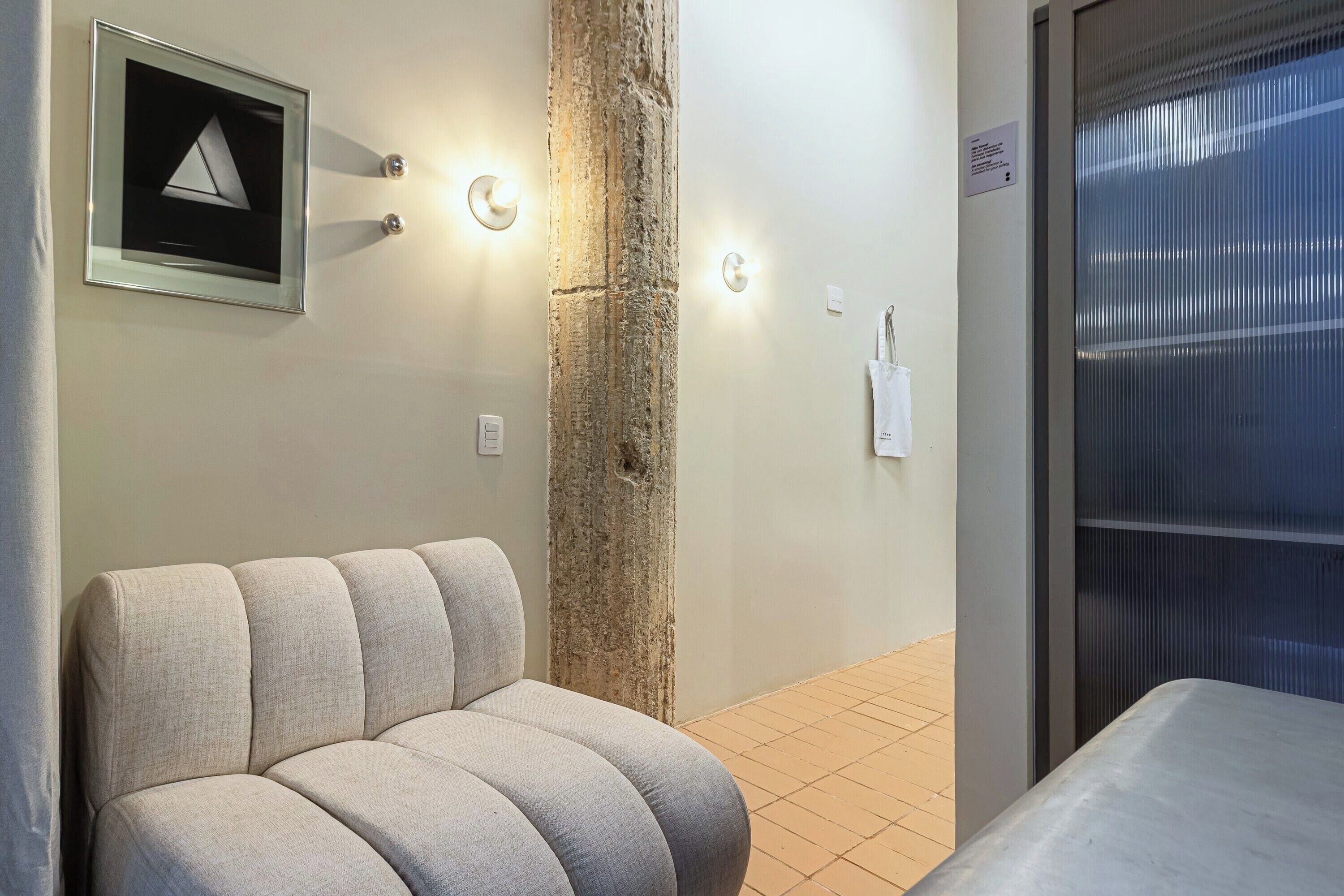 A hotel room with a sofa, a pillar, wall-mounted lights, a framed picture, and a window with blinds.