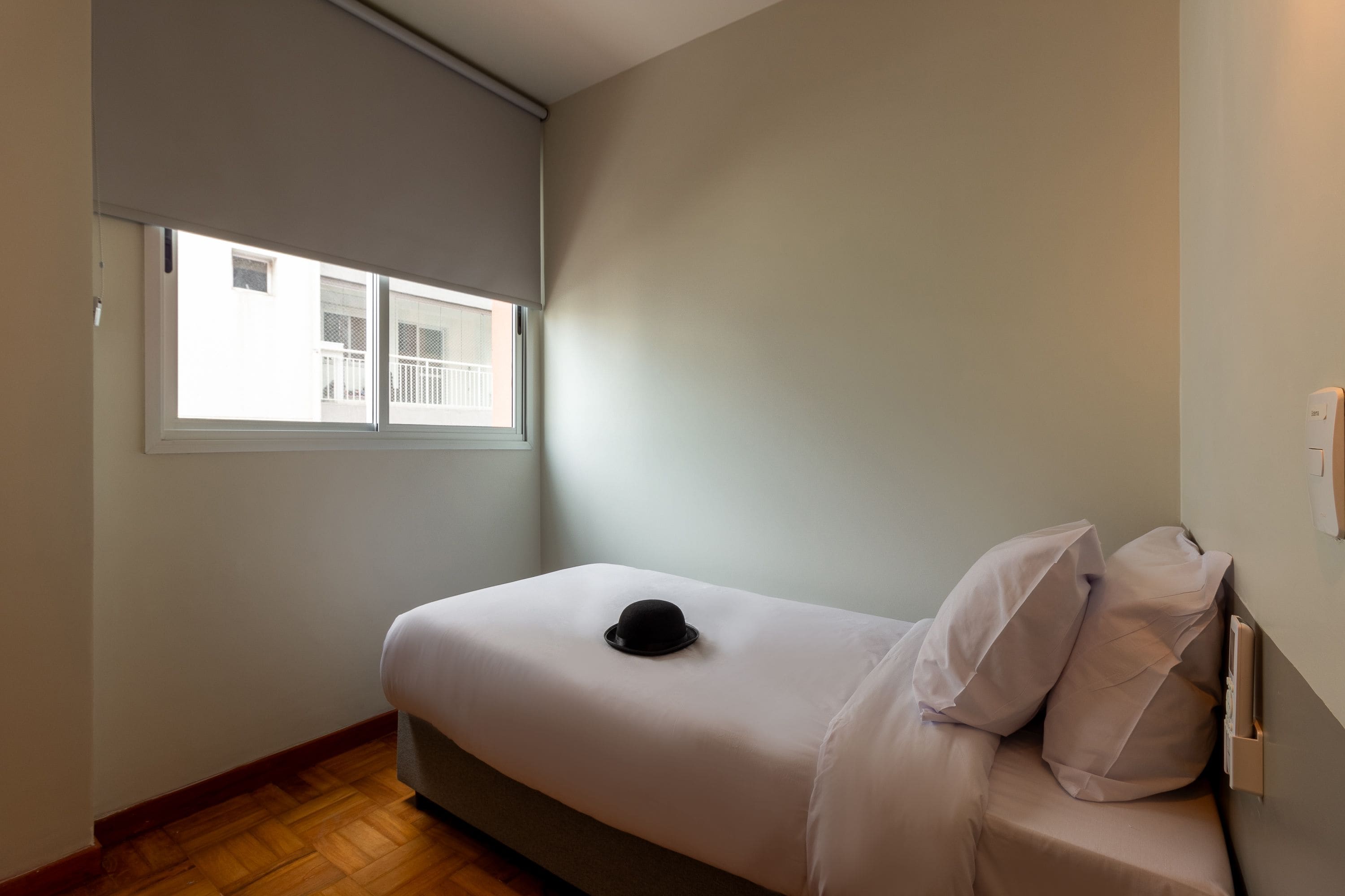 A small, single-bed room with a window, a wall-mounted thermostat, and a wooden floor.