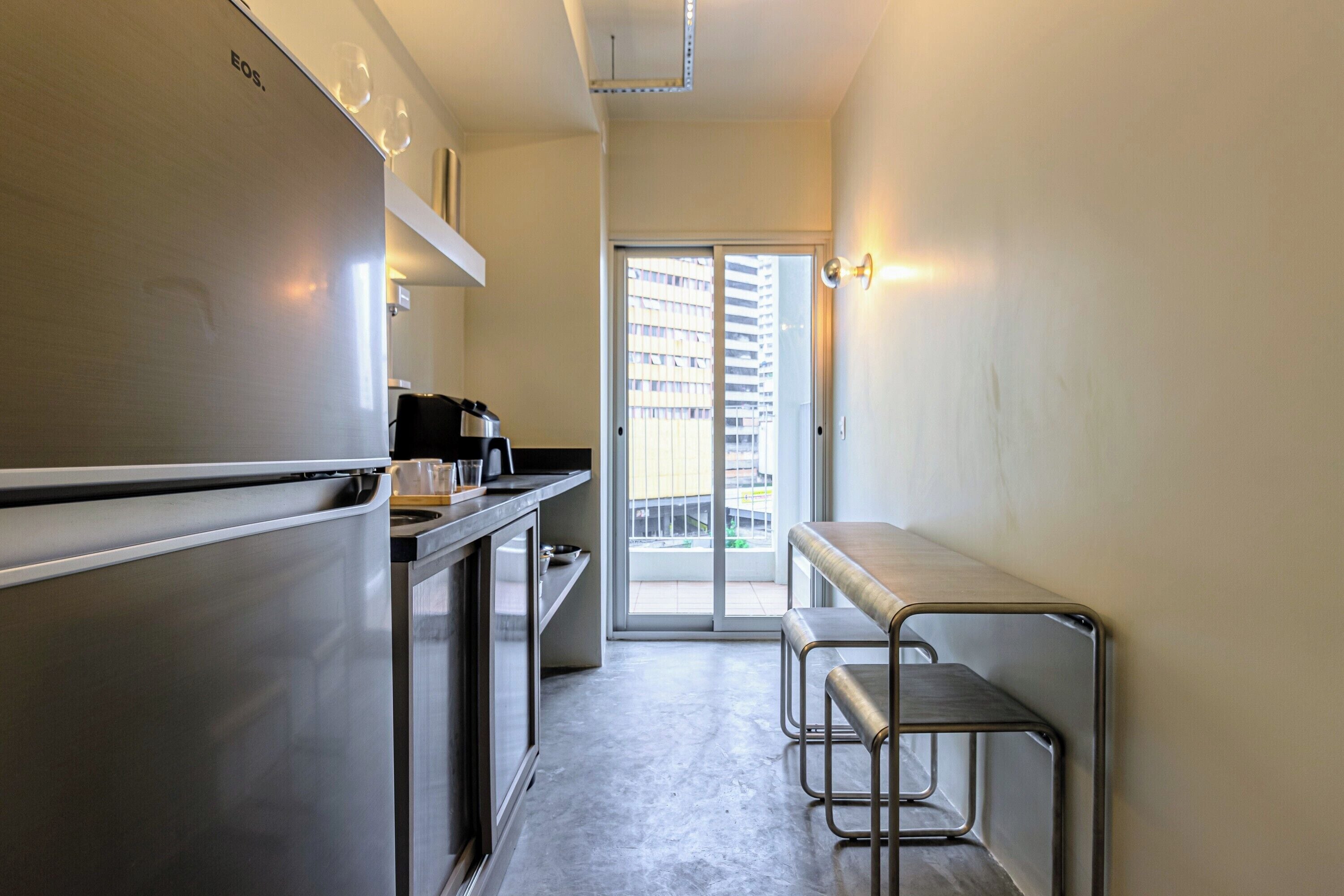 Une cuisine étroite équipée d’un réfrigérateur en acier inoxydable, d’une machine à café et d’un petit plan de travail. Une porte-fenêtre coulissante donne accès à un balcon d’où l’on peut admirer la vue sur les immeubles.