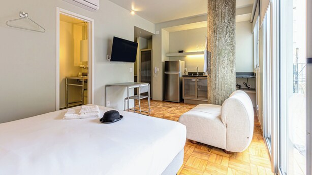 A hotel room with a bed, a chair, a desk, a television, and a view of the kitchen and outside. - Charlie Petrah Vila Buarque - Soft Opening (São Paulo)