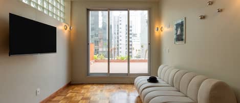 A modern living room with a flat-screen TV mounted on the wall, a beige sofa, and a large window with a view of buildings and greenery.