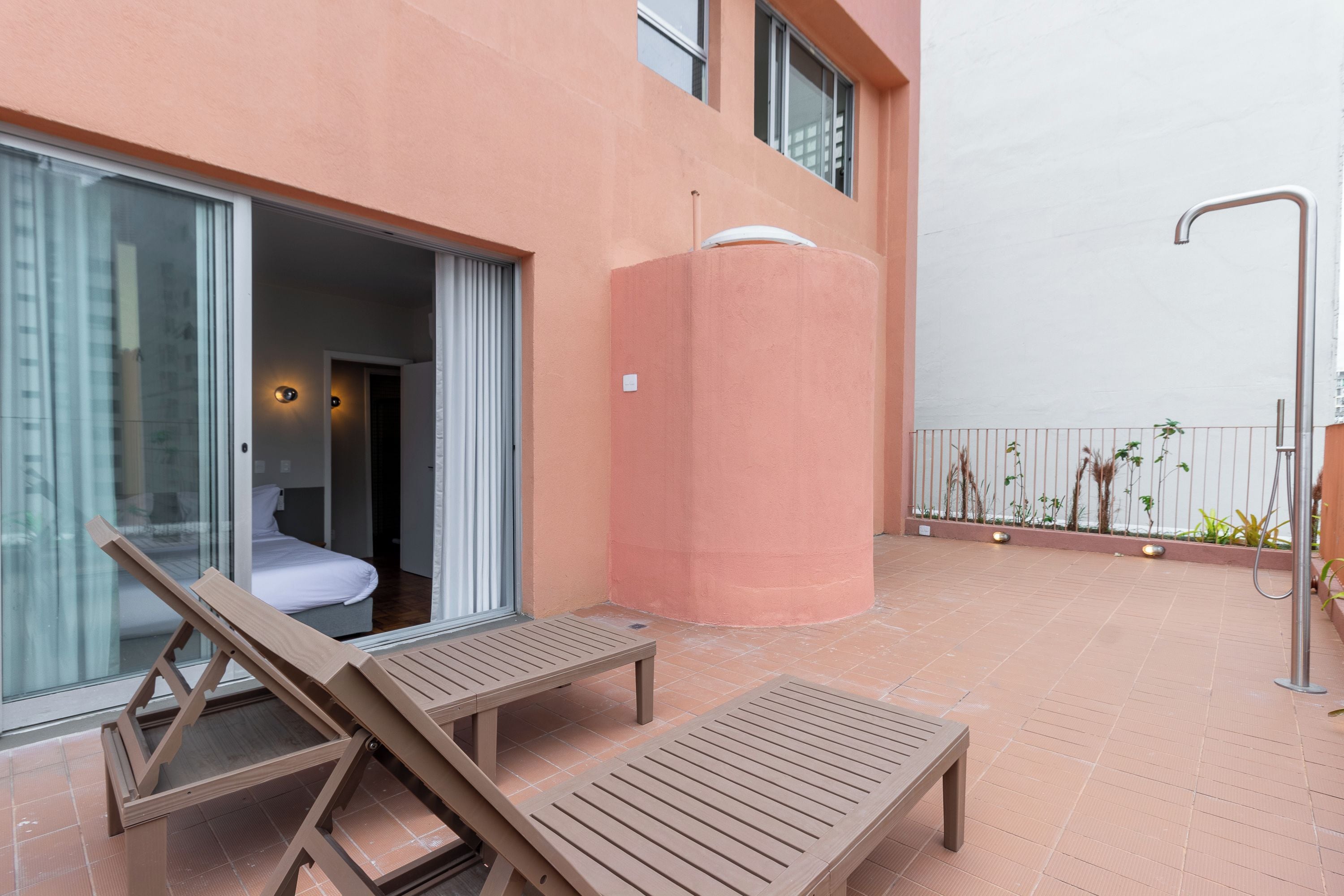 A balcony with a wooden lounge chair, a second lounge chair, a glass door leading to a room with a bed, and a water feature.