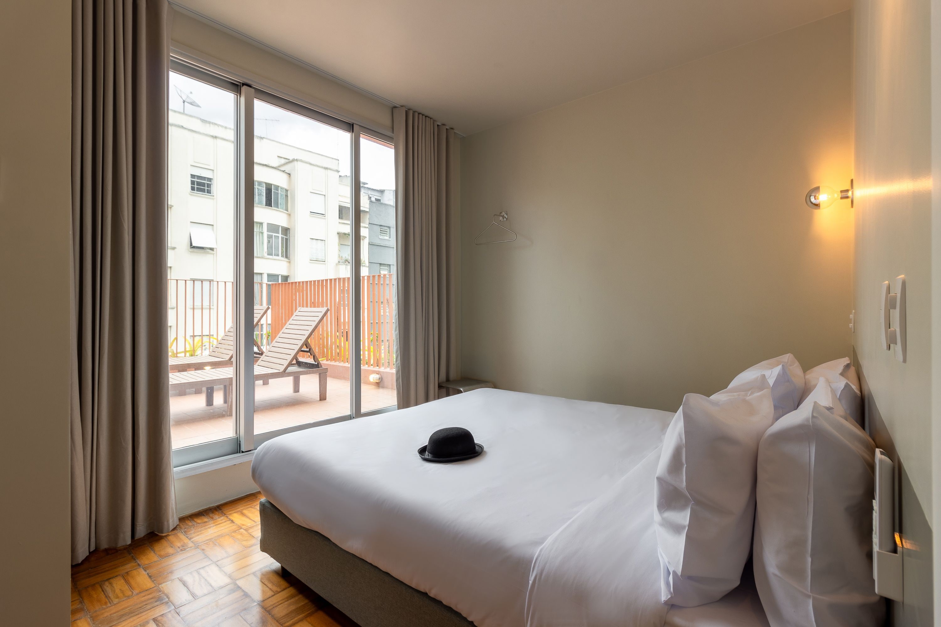 A hotel room with a bed, a hat on the bed, a window with curtains, and a view of the outside balcony.