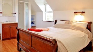 A bedroom with a wooden bed, a nightstand, a mirror, a window, and a bathroom with a sink and cabinets. - 4 Bed in Caherdaniel (oc-ii04851) (Caherdaniel)