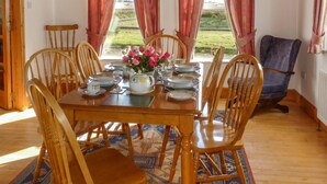 A dining area with a wooden table set for four, surrounded by wooden chairs, with a view of a garden through large windows. - 3 Bed in Achill Island (oc-ii06848) (Doogort)