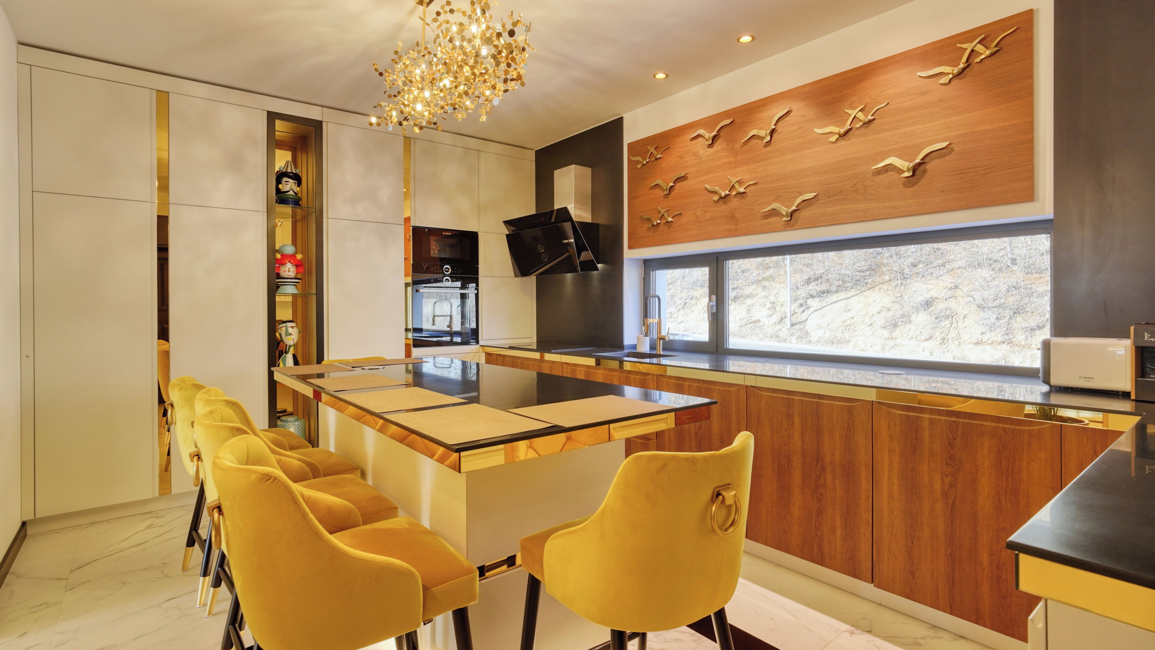 Une cuisine moderne avec un îlot central, des chaises jaunes, un mur en bois décoré de motifs d’oiseaux et une grande fenêtre.