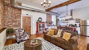 Un vaste salon avec un mur en briques, un plafond en bois et une cuisine moderne en arrière-plan.