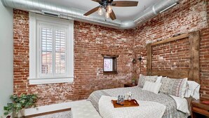 Une chambre avec des murs en briques apparentes, un lit avec une literie à motifs, un ventilateur de plafond et une fenêtre avec des stores.