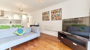 Ein modernes Wohnzimmer mit einer Couch, einem Flachbildfernseher, Holzböden und einer Küche im Hintergrund.