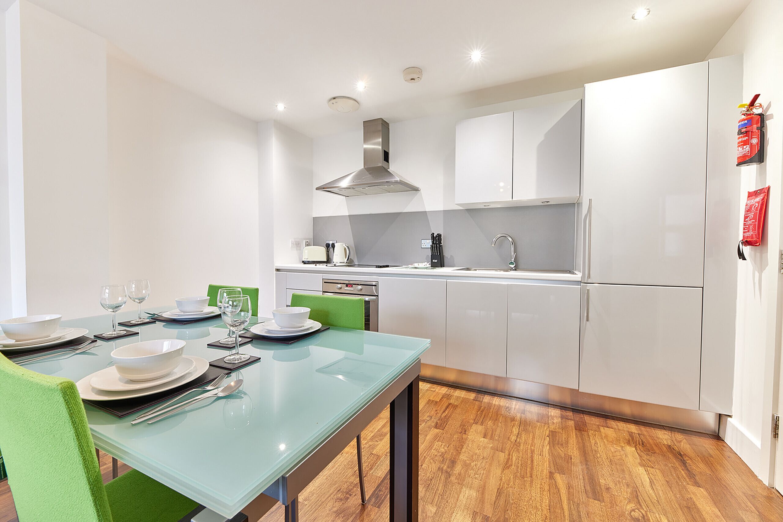 Een moderne keuken met witte kastjes, een roestvrijstalen afzuigkap, en een eetgedeelte met een glazen tafel en groene stoelen.