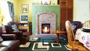 A living room with a brick fireplace, leather sofas, a dining table with a fruit-patterned tablecloth, and a television on a stand. - 1 Bed in Ballysadare (oc-ii14644) (Drumiskabole West)