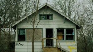 A dilapidated house with boarded-up windows, a wooden staircase, and graffiti on the side.