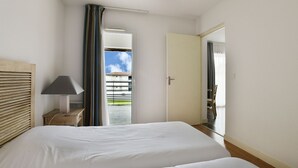 Ein Hotelzimmer mit einem großen Bett, einem Nachttisch mit Lampe, Blick auf einen Außenbalkon und einer Tür zu einem anderen Raum.