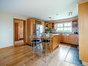 A modern kitchen with wooden cabinets, a stainless steel refrigerator, a dining area with two bar stools, and a window providing natural light.