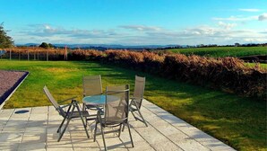 Een terras buiten met een tafel en stoelen, een stenen vloer en uitzicht op een grasveld en heuvels in de verte.