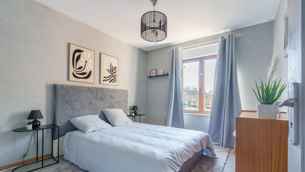 Ein Schlafzimmer mit Bett, Nachttischen, einer Lampe, einer Deckenleuchte, einem Fenster mit Vorhängen und einer Zimmerpflanze.