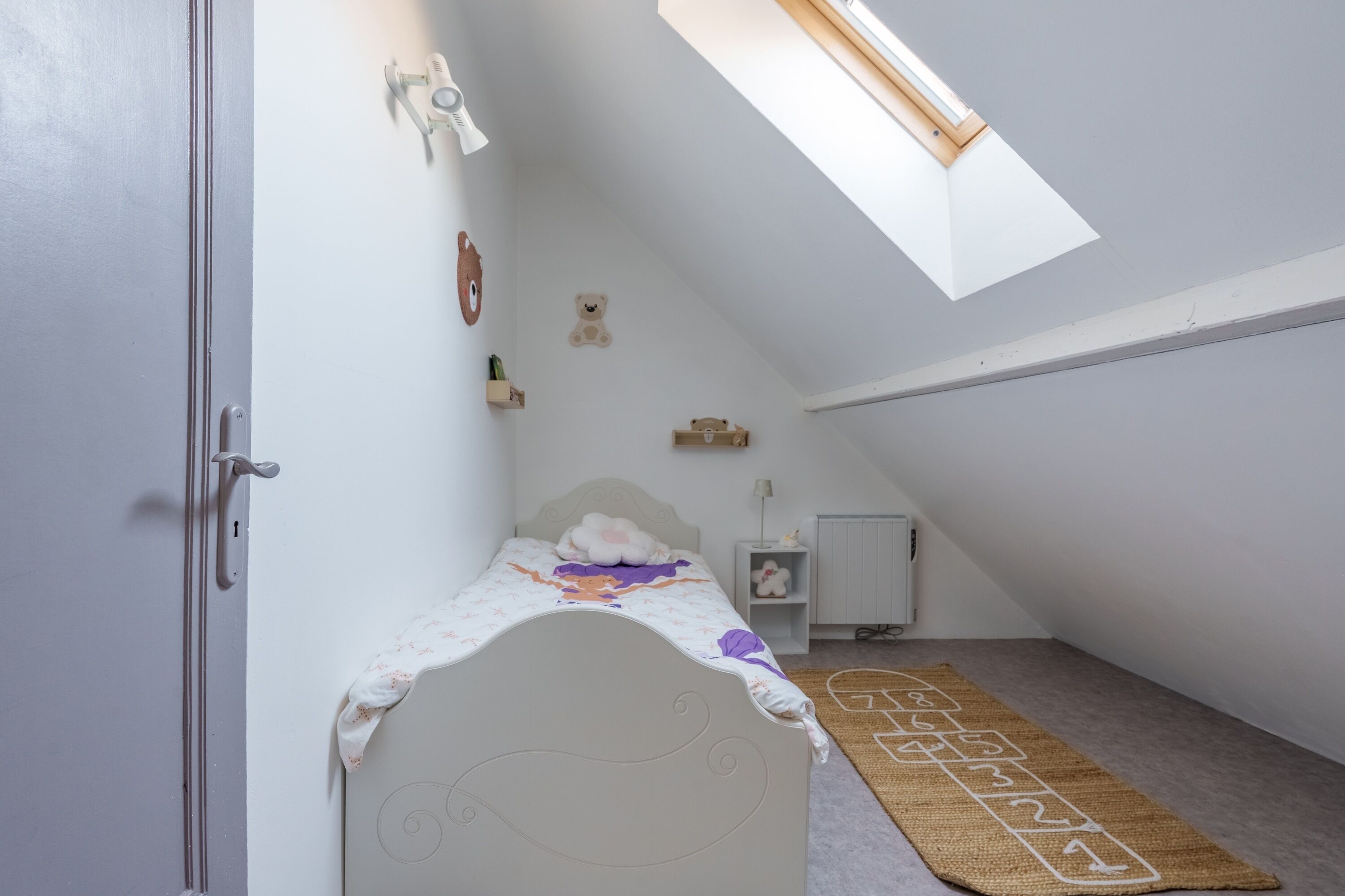 Quarto com teto inclinado, cama, radiador, prateleira pequena e janela.