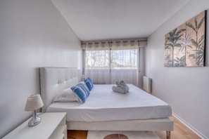 Ein Schlafzimmer mit einem Bett, einem Nachttisch, einer Lampe, einem Fenster mit Vorhängen und einem Wandbild.