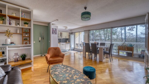 A spacious living room with a leather sofa, a patterned ottoman, a bookshelf, and a dining area with a table and chairs. - Le Halo - Avec Balcon et Parking Privé (Le Bouscat)