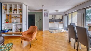 A modern living room with a wooden floor, a pink chair, a dining area with a table and chairs, a kitchen in the background, and a large window. - Le Halo - Avec Balcon et Parking Privé (Le Bouscat)