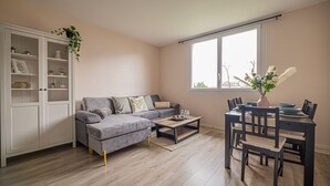 Una sala de estar con un sofá gris, una mesa de comedor con sillas, un armario blanco con puertas de vidrio y una ventana con vista a árboles.