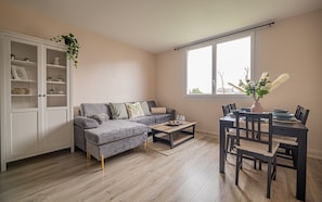 A living room with a grey sofa, a dining table with chairs, a white cabinet with glass doors, and a window with a view of trees. - Hesychia - A 1 km du centre historique (Le Mans)