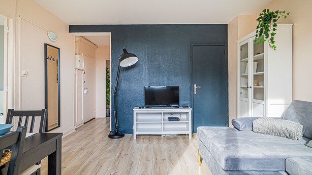 Una sala de estar con una televisión sobre un mueble blanco, un sofá, una mesa de comedor con sillas y una lámpara.