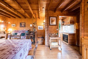 Interior de una cabaña de madera con una cama, una mesa, una silla, una zona de cocina y cuadros en las paredes.
